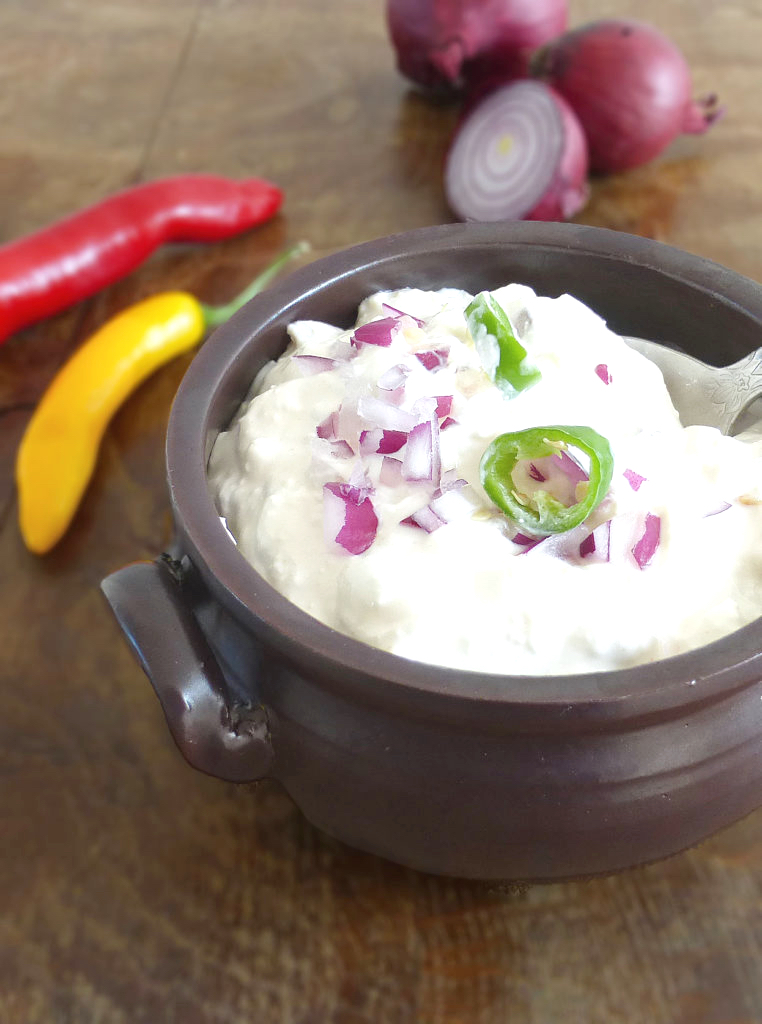 a brown bowl filled with yoghurt dip topped with red onion and green chillies