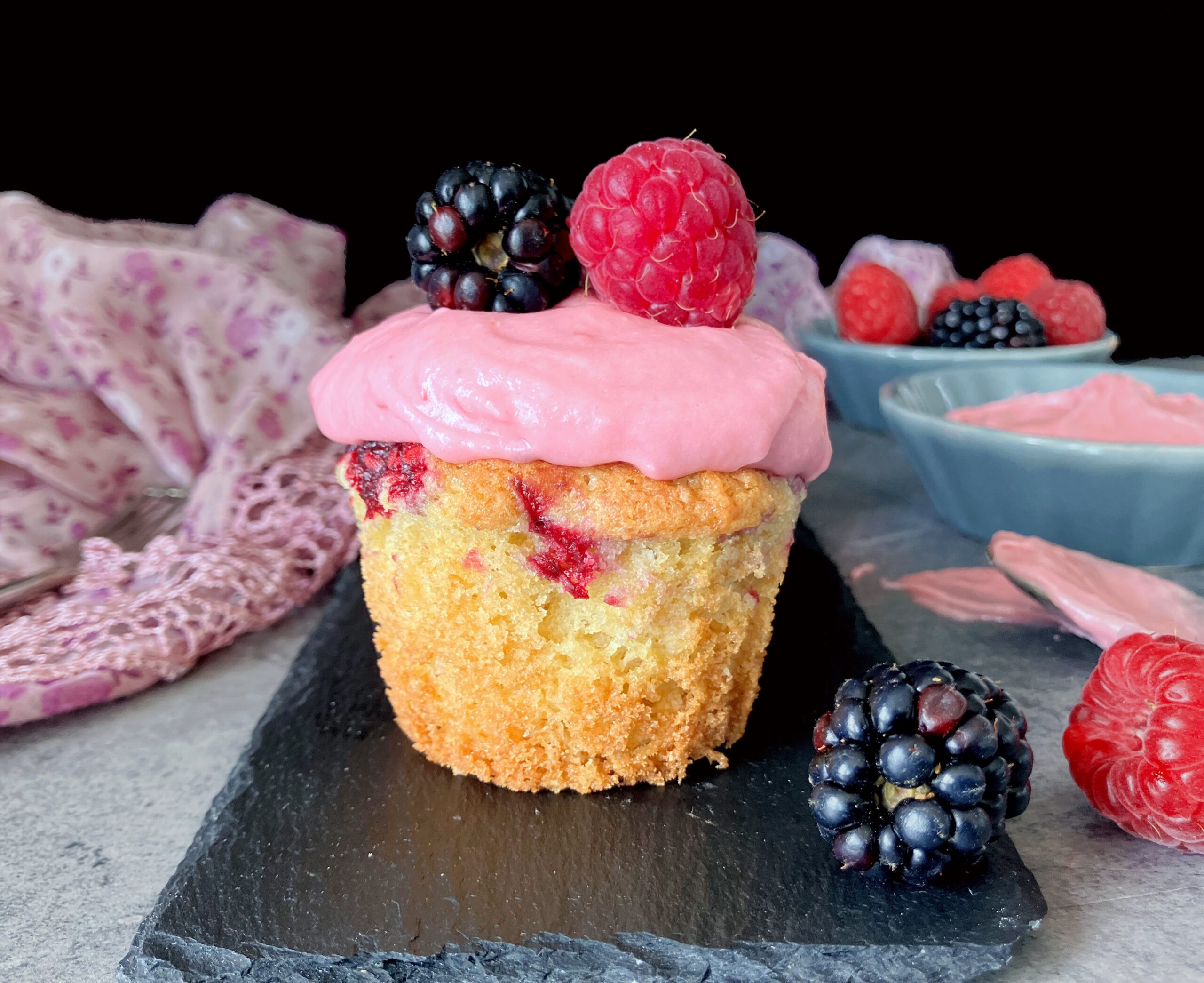 Cupcake with pink frosting topped with berries on a black slate with a pink cloth