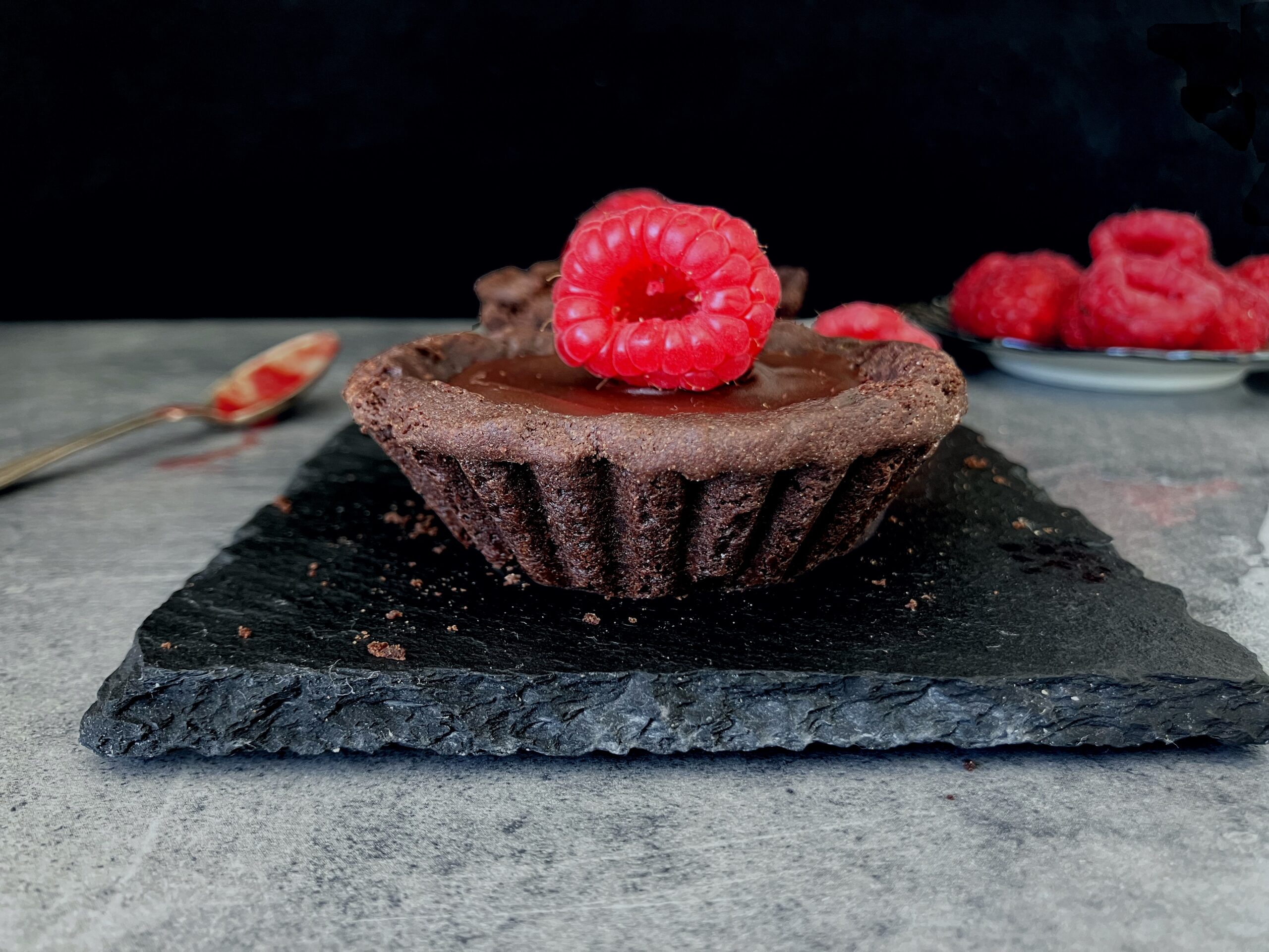 dark chocolate tartlet on slate with raspberry on top and a black background